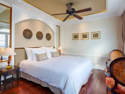 Ein stilvolles Schlafzimmer an Bord des Flusskreuzfahrtschiffs Mekong Navigator. Zu sehen sind ein großes Bett, Nachttisch, Lampe, zwei Holzsessel und eine Decke mit Ventilator.