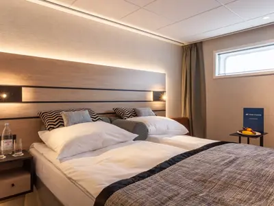 Geräumige Doppelkabine an Bord des Flusskreuzfahrtschiffs nickoSPIRIT mit zwei Einzelbetten, Holz-Kopfteil, Leselampen und einem Fenster. Auf einem Tisch liegt ein Obstteller bereit.
