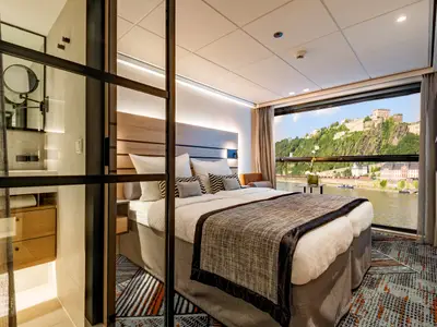 Kabine des Flusskreuzfahrtschiffs nickoSPIRIT: Doppelbett, Panoramafenster mit Flussblick, bewaldete Hügel und Festung. Badbereich hinter Glastrennwand sichtbar.