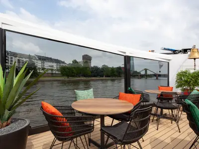 Das Außendeck der nickoSPIRIT zeigt Tische, Stühle mit bunten Kissen und Pflanzen. Die Fenster reflektieren die Frankfurter Flusslandschaft mit Gebäuden und einer Brücke.