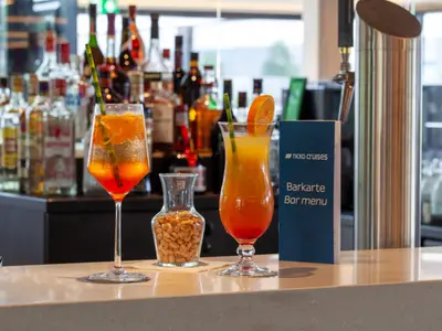 Zwei bunte Cocktails stehen auf einer hellen Bartheke. Hinten eine gut bestückte Bar mit Flaschen und einer Barkarte mit "Nicko Cruises" Logo.