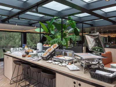 Frühstücksbuffet auf der nickoSPIRIT. Es gibt warme Speisen, Brot, Aufschnitt und Getränke. Große Fenster zeigen Fluss und Ufer. Die helle Kabine hat ein Glasdach und moderne Einrichtung.
