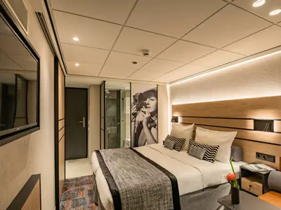 Moderne Kabine der nickoSPIRIT: Doppelbett, Holzkopfteil mit Beleuchtung, Wand-TV. Glasdusche und Kunstfotografie sichtbar.