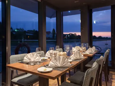 Abendliche Szene im Restaurant der nickoSPIRIT. Gedeckte Tische, weiße Servietten, Gläser. Große Fenster bieten Blick auf Fluss und Dämmerung mit Ufer.