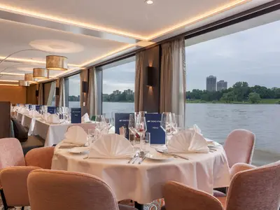 Blick in das modern eingerichtete Restaurant der nickoSPIRIT. Elegante Tische sind gedeckt. Große Fenster bieten Flussblick auf Bäume und Gebäude am Ufer.