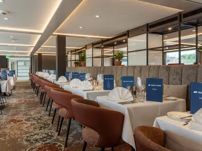 Das helle Restaurant der nickoSPIRIT zeigt gedeckte Tische mit weißen Tischdecken, Stühlen und blauen Menüs. Eine gepolsterte Sitzbank und Fenster mit Ausblick sind ebenfalls zu sehen. Moderne Beleuchtung und ein gemusterter Teppichboden prägen den Essbereich auf der nickoSPIRIT.