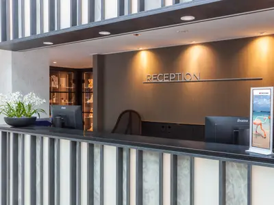 Moderne Rezeption mit dunkler Theke und vertikalen Designelementen. Ein Blumenarrangement steht links, rechts ein Digital-Display mit Kreuzfahrtwerbung. Im Hintergrund sind beleuchtete Shop-Vitrinen zu sehen, auf einem Display "SHOP" ist lesbar.