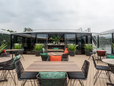 Das helle Sonnendeck der nickoSPIRIT zeigt Tische, bequeme Stühle und große Topfpflanzen. Dahinter ein verglaster Innenbereich mit Buffet und Flussblick.