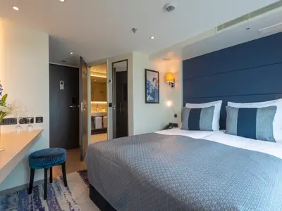 Modernes Schlafzimmer der nickoVISION: Doppelbett mit blauer Kopflehne, Leselampen und Schreibtisch. Eingang zum Bad im Hintergrund sichtbar.