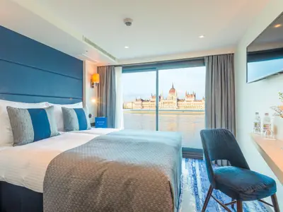 Innenansicht einer modernen Kabine des Flusskreuzfahrtschiffs nickoVISION mit Doppelbett und Blick auf das ungarische Parlament am Donauufer.