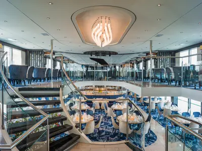 Blick in das zweistöckige Restaurant der nickoVISION mit geschwungener Treppe, Tischen, Stühlen und einem Kronleuchter.