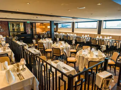 Restaurant an Bord der nickoVISION. Tische mit weißen Tischdecken und Gedecken auf zwei Ebenen, durch Geländer getrennt. Fenster und Sitzbank im Hintergrund.