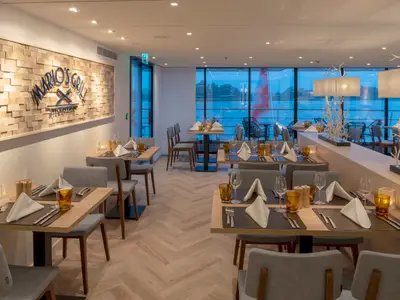 Das Restaurant Mario's Grill auf dem Flusskreuzfahrtschiff nickoVISION. Man sieht stilvoll gedeckte Tische, eine Holzwand mit Restaurantlogo und große Fenster mit Flussblick.