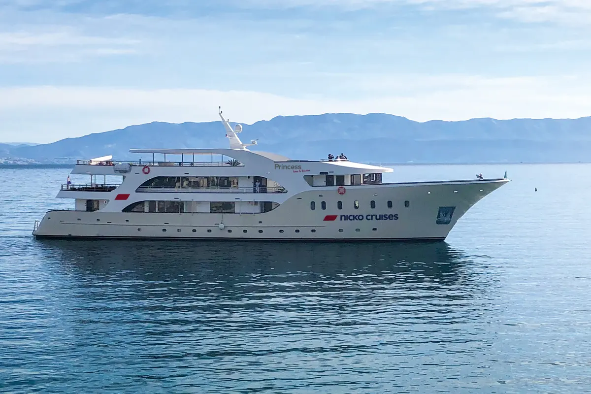 Das weiße Kreuzfahrtschiff 'Princess' von Nicko Cruises fährt auf ruhigem Meer. Im Hintergrund Berge unter blauem Himmel. Mehrere Decks mit Fenstern und wenige Passagiere sind sichtbar.