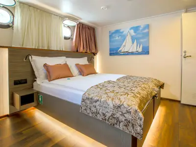 Kabine der nicko cruises Princess mit Doppelbett, Leselampen, zwei Bullaugen mit Vorhängen, einem Segelboot-Gemälde und einer weißen Tür.