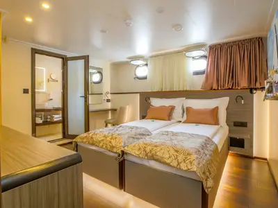 Kabine des Flusskreuzfahrtschiffs Princess mit zwei Betten, Porthole-Fenstern, Schreibtisch und Bad. Warme Beleuchtung unterstreicht die Holzeinrichtung.