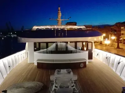 Das Vordeck der Princess bei Nacht. Beleuchtetes Teakholzdeck, weiße Relings, Mast und helle Hafenpromenade mit Palmen. Links dunkles Hafenwasser.