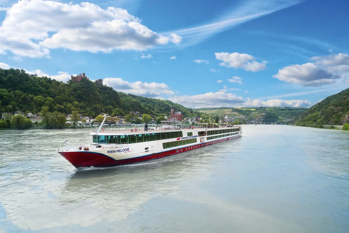 Das weiß-rote Flusskreuzfahrtschiff "Rhein Melodie" fährt auf dem Rhein. Grüne Hügel mit Burgruine, Kirche und Weinbergen säumen das Ufer unter blauem Himmel.
