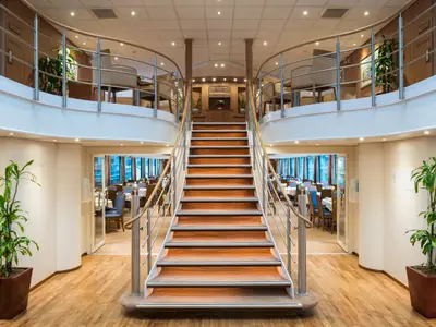 Innenansicht des Flusskreuzfahrtschiffs Rhein Melodie. Eine große Holztreppe in der Mitte führt zu einer Galerie. Links und rechts sind Speisesäle zu sehen.