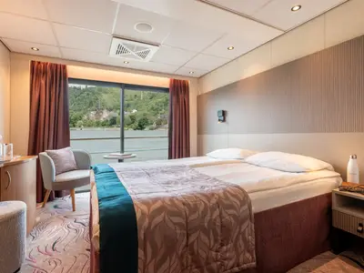 Helle Kabine der Rhein Melodie mit Panoramafenster. Blick auf Fluss und grüne Landschaft mit Dorf. Doppelbett, Sessel und Schreibtisch sind erkennbar.