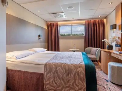 Blick in eine Kabine der Rhein Melodie: Ein gemütliches Doppelzimmer mit breitem Bett, Fenster, Sessel und Arbeitsplatz mit TV. Helle, moderne Einrichtung mit Teppichboden.