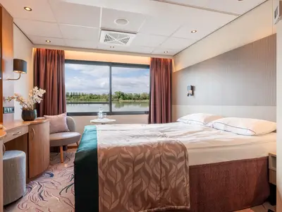 Moderne Kabine auf der Rhein Melodie mit Bett, Schreibtisch, Sessel und Panoramafenster. Draußen sind ein Fluss und bewaldete Ufer sichtbar.