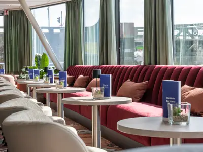 Die Lounge der Rhein Melodie zeigt rote Sitzbänke und helle Stühle. Runde Tische mit Barkarten und Dekoration stehen vor einer durchgehenden Fensterfront mit Ausblick.