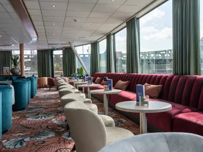 Blick in die helle Lounge der Rhein Melodie: Rote Samtbänke, helle Sessel und türkise Hocker auf einem Teppich. Große Fenster zeigen die Flusslandschaft und eine Brücke.