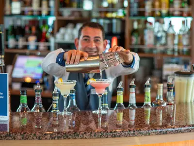 Ein lächelnder Barkeeper auf der Rhein Melodie schenkt einen Cocktail aus einem Shaker in ein Glas. Auf der Theke stehen weitere fertige Drinks und Flaschen bereit.