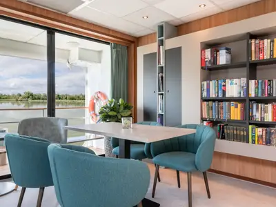 Rhein Melodie Lounge: Türkise Sessel, ein Tisch, Bücherregale. Fenster mit Blick auf Fluss, bewaldetes Ufer, Rettungsring.