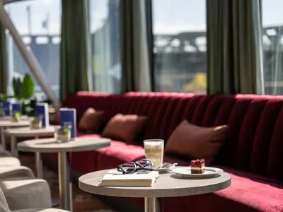 Eine helle, moderne Lounge mit einem langen, roten Samtsofa und braunen Kissen entlang einer Fensterfront. Mehrere kleine Tische stehen davor. Auf dem vordersten Tisch liegen eine Sonnenbrille, ein Buch, ein Latte Macchiato und ein Stück Kuchen. Tageslicht flutet herein.