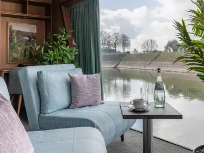 Einladende Kabine der Rhein Melodie mit hellblauen Polstermöbeln, einem Tisch mit Kaffee und Wasser. Durch das große Fenster ist ein Flussufer mit Bäumen und bewölktem Himmel zu sehen.
