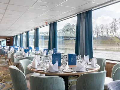 Das helle Restaurant des Flusskreuzfahrtschiffs Rhein Melodie zeigt mehrere gedeckte Tische mit teal-farbenen Stühlen. Große Fenster bieten einen Ausblick auf einen Fluss, eine grüne Brücke und das Ufer.