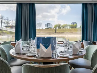 Elegantes Flusskreuzfahrt-Restaurant mit gedeckten Tischen und Panoramablick auf den Fluss und die Uferlandschaft. Keine Personen sichtbar.