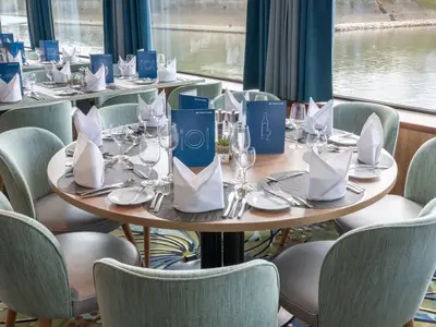 Restaurant an Bord der Rhein Melodie mit gedeckten Tischen, Stühlen und blauen Menükarten. Große Fenster zeigen den Fluss. Viel Licht.
