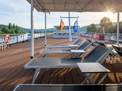 Blick auf das Sonnendeck der Rhein Melodie mit Liegestühlen. Der Fluss und bewaldete Hügel sind im Hintergrund. Eine deutsche und nicko cruises Flagge wehen.