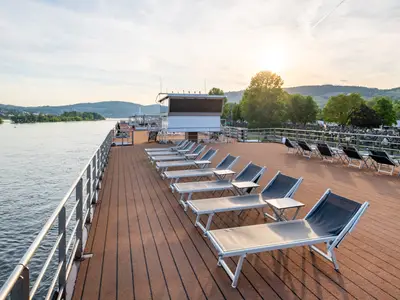 Das Sonnendeck der Rhein Melodie bei Sonnenuntergang. Eine Reihe leerer Liegestühle mit Tischen steht bereit. Links fließt der Fluss, rechts sind grüne Hügel zu sehen.