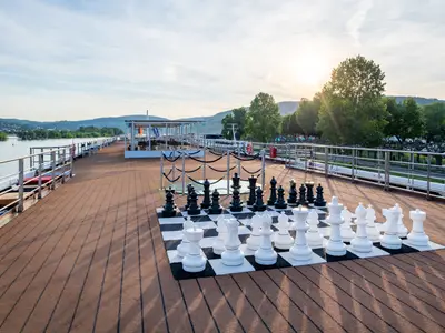 Ansicht des Sonnendecks der Rhein Melodie mit großem Schachspiel, Lounge-Bereichen, Fluss und bewaldeten Hügeln bei Sonnenlicht.