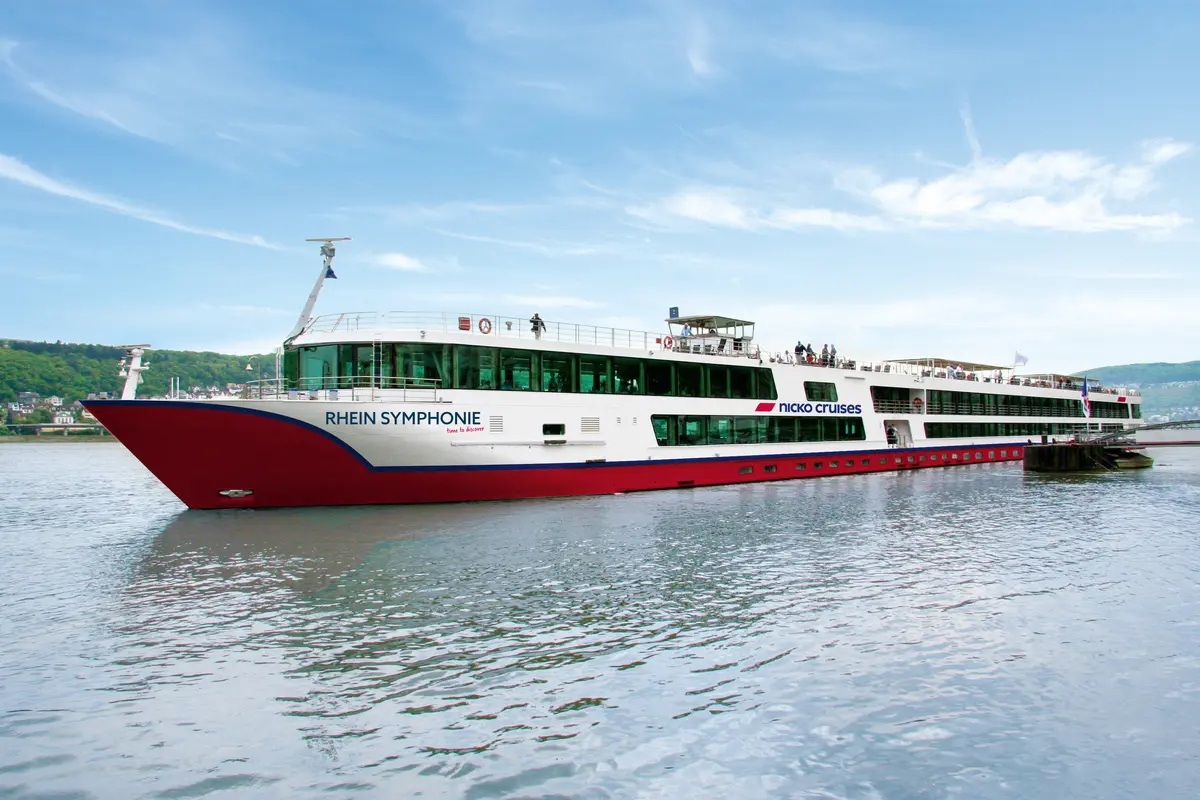Das Flusskreuzfahrtschiff MS Rhein Symphonie von nicko cruises fährt auf dem Wasser. Es hat einen roten Rumpf und weiße Aufbauten mit vielen Fenstern. Mehrere Passagiere sind auf den Außendecks zu sehen. Im Hintergrund sind grüne Hügel und eine kleine Ortschaft.