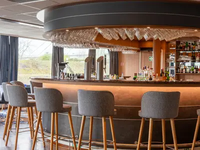 Innenansicht der Bar auf der Rhein Symphonie. Eine beleuchtete Theke mit Spirituosen, Gläsern und Barhockern. Große Fenster mit Blick nach draußen sind sichtbar.