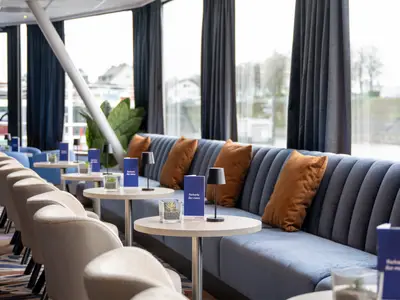 Eine moderne Bar-Lounge mit blauen, geriffelten Sofas, orangefarbenen Kissen und kleinen Tischen. Auf den Tischen stehen schwarze Lampen, Pflanzen und blaue Barkarten. Große Fenster geben den Blick auf eine Flusslandschaft frei.