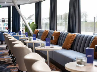 Die Lounge der Rhein Symphonie präsentiert sich mit blauen Sitzbänken, orangen Kissen und hellen Sesseln. Kleine Tische mit Bar-Menüs und Lampen. Große Fensterfront zum Fluss.