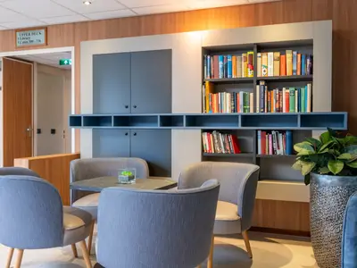 Eine Lounge auf der Rhein Symphonie. Man sieht bequeme Sessel um kleine Tische, gefüllte Bücherregale und eine Wegbeschreibung zum Oberdeck und zur Bibliothek.