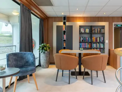 Ein gemütlicher Lounge- und Bibliotheksbereich an Bord der Rhein Symphonie. Bequeme Sessel, Tische, voll bestückte Bücherregale und große Fenster mit Blick auf den Fluss sind sichtbar.