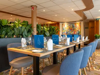 Restaurant der Rhein Symphonie: Gedeckte Tische mit blauen Menükarten und Stühlen in Blau und Grau. Eine üppige grüne Pflanzwand und ein farbenfrohes Gemälde schmücken den Raum.