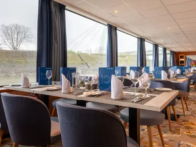 Restaurant der Rhein Symphonie: Gedeckte Tische an Fenstern mit winterlicher Flusslandschaft im Hintergrund. Blaue Menüs, graue Stühle. Hinten Bar und Spiegel.