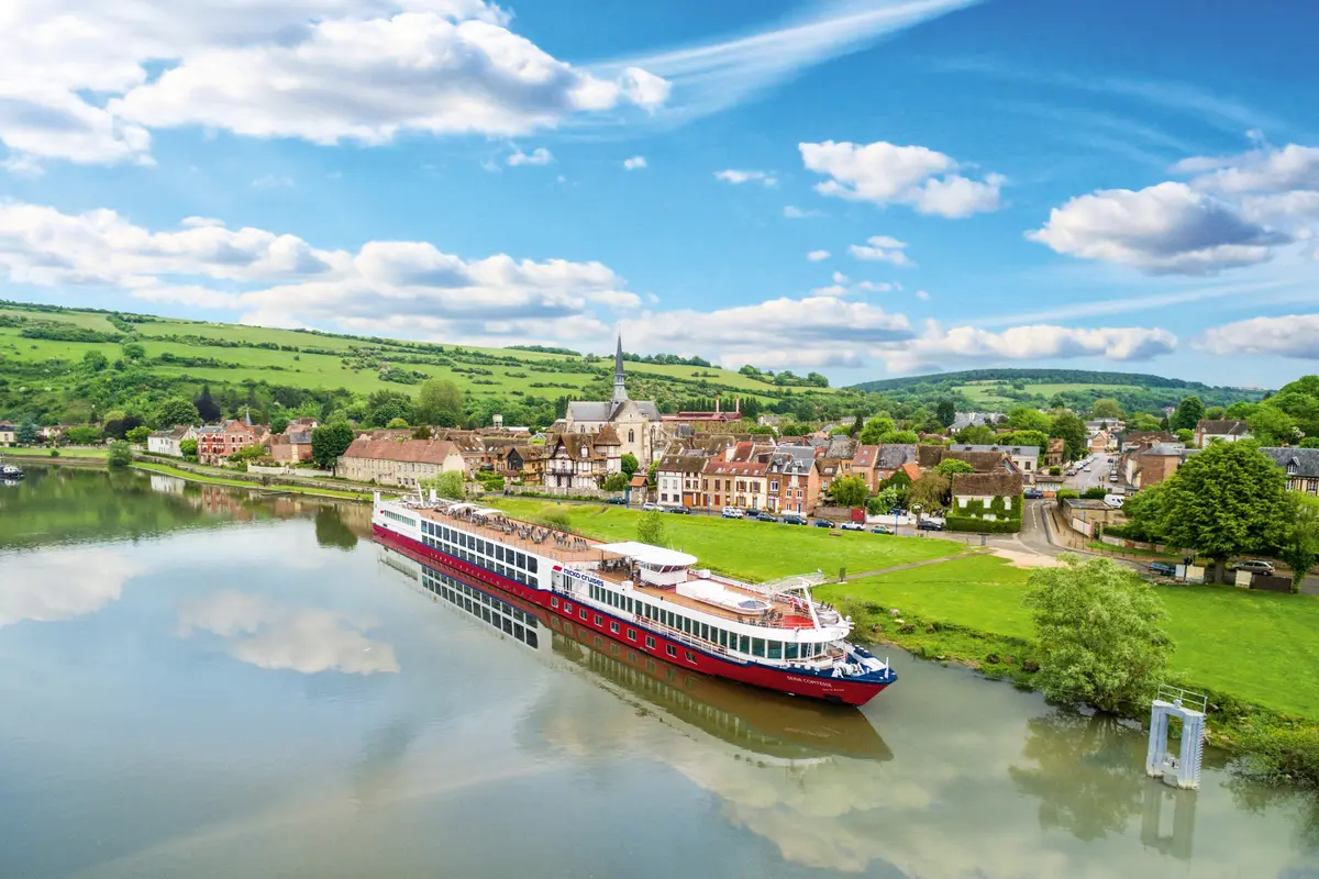 Die 'Seine Comtesse', ein rotes Flusskreuzfahrtschiff, liegt auf dem Fluss. Eine malerische Stadt mit Kirche und grünen Hügeln unter blauem Himmel bildet die Kulisse.