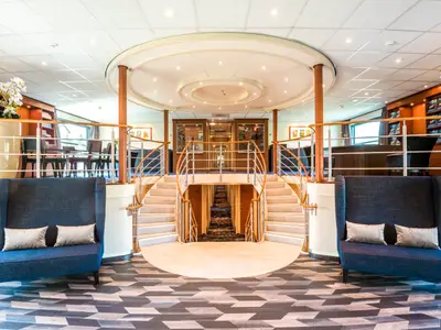 Die Lobby der Seine Comtesse. Zentrale Doppeltreppe. Blaue Sessel im Vordergrund auf geometrischem Boden. Seitlich sind Loungebereiche mit Tischen und Stühlen sichtbar.