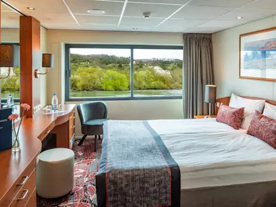 Kabine der Seine Comtesse. Doppelbett, großes Fenster mit Flussblick. Schreibtisch, Sessel und nicko cruises Infomappe.