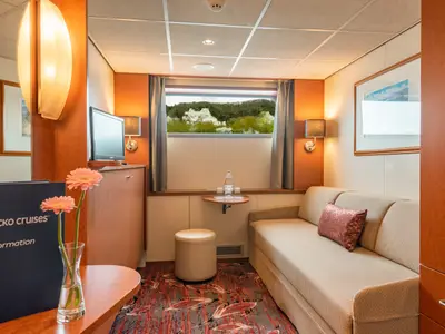 Innenansicht einer hellen Kabine an Bord der Seine Comtesse. Man sieht ein beiges Sofa, einen Tisch mit Blumen und Fensterblick auf eine grüne Landschaft. Zur Ausstattung gehören TV und Wandlampen.